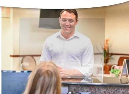 A man stands behind a countertop barrier at reception desk to greet a woman